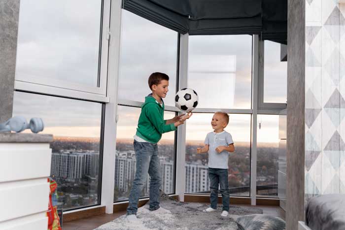 Two young children playing with a soccer ball inside a high-rise apartment near large floor-to-ceiling windows overlooking the city, enjoying natural light and safety while showcasing comfort and professional Window Replacement Marietta services.