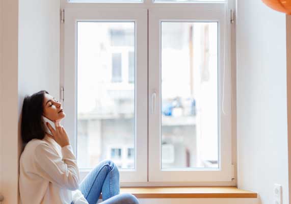 Woman sitting by a white-framed window talking on the phone in a bright modern room, enjoying natural light and city views while highlighting comfort, style, and professional Window Replacement Marietta services.