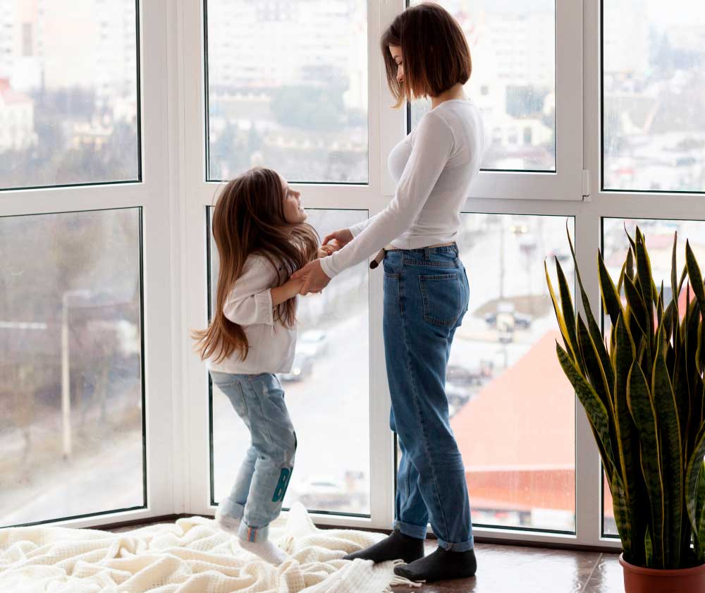 Mother and young daughter standing by large floor-to-ceiling windows in a bright apartment, enjoying natural light and city views while highlighting family comfort, safety, and professionally installed New & Replacement Windows Atlanta.