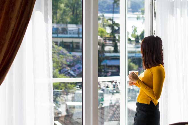 Woman standing by large bright windows holding a cup and looking outside, enjoying natural light and city views in a modern home, showcasing comfort, style, and professionally installed New & Replacement Windows Atlanta.